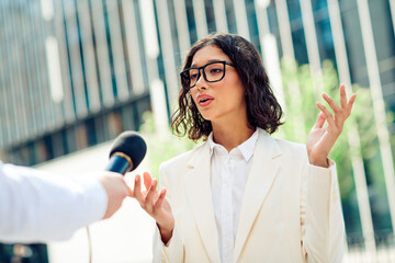 Successful businesswoman giving an interview outdoors against a modern office building, dressed in...
