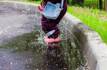 The child jumps through the puddles. The child in a bright cloak and colorful rubber boots joyfully...