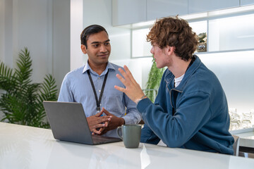 Two men engaged in a discussion at a modern kitchen table, with a laptop and a coffee cup present,...