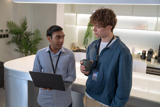 Two men engage in a discussion while looking at a laptop in a modern office setting, with coffee and greenery in the background.
