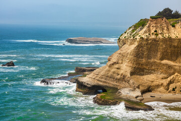 Waves crash against sculpted sandstone cliffs and sea caves along the rugged Pacific Coast near...