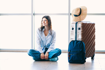 Young female tourist sitting with luggage in modern airport terminal, traveling in casual style for...