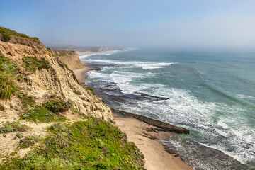Waves crash against sculpted sandstone cliffs and sea caves along the rugged Pacific Coast near...