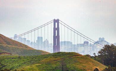 Golden Gate Bridge tower framed by lush hills of Marin Headlands with San Francisco skyline in the background seen through coastal haze on a spring day