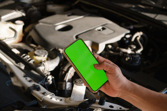 Hand holding a smartphone with green screen in front of an open car hood, representing digital roadside assistance.
