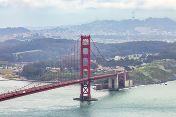 Panoramic view of the Golden Gate Bridge spanning San Francisco Bay with cityscape, lush hills, and boats, captured from a high vantage point on a cloudy spring day