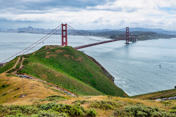 Panoramic view of the Golden Gate Bridge spanning San Francisco Bay with cityscape, lush hills, and boats, captured from a high vantage point on a cloudy spring day