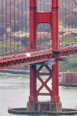 Close-up of Golden Gate Bridge tower with Palace of Fine Arts in the background, San Francisco cityscape and boats on the bay visible through the iconic red structure.