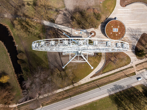 Aerial perspective of a Ferris wheel, circular building with green roof, pathways, parking area, road, grass, trees, and a small waterway.