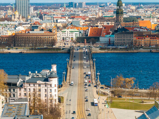 A bridge spans a wide river in a bustling city, with vehicles on the road, a white turreted building, colorful rooftops, and a church with a green spire.