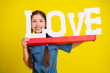 Smiling girl holding a large love sign with a vibrant yellow background expressing happiness and...