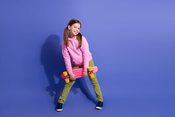 Adorable young girl in a pink hoodie holding a skateboard, posing confidently against a vibrant purple background