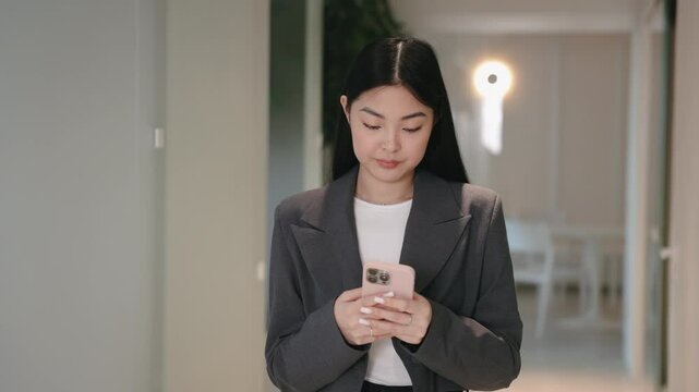 Focused Asian woman in formal blazer looking down at smartphone and typing with both thumbs. Sending email or replying to message. Staying concentrated while managing digital tasks in hallway.