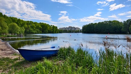 A plastic boat is moored on the lake shore next to a sandy beach and reeds. On the far shore are...