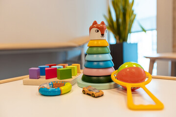 Obraz premium A variety of children's toys, including a fox stacking toy, yellow car, rattle, and blocks, arranged on a white table with a potted plant visible.