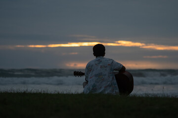 A young man sits and plays guitar on a tropical beach at sunset. A happy man enjoys the sunset while playing guitar on the beach