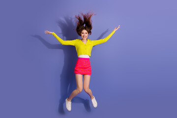Joyful young woman jumping against a vibrant purple background wearing colorful outfit and...