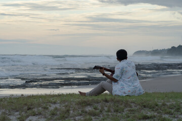 A young man sits and plays guitar on a tropical beach at sunset. A happy man enjoys the sunset while playing guitar on the beach