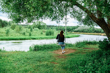 A happy young girl is sitting on a swing in a beautiful tree by the river.