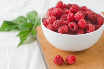 Ripe pink raspberries in a bowl with green leaves on a white background. Concept of summer berry harvest. Horizontal orientation. Selective focus.