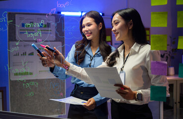 Two businesswomen working late at night, planning a business project on a transparent glass board in a modern office