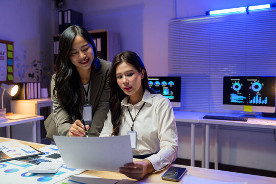 Two Asian businesswomen reviewing marketing reports, analyzing financial statistics, and discussing investment strategies in a brightly lit office at night