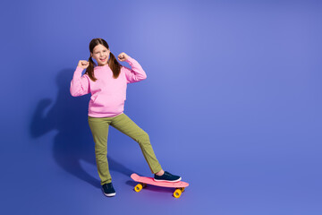 Cheerful young girl with a pink skateboard posing against a vibrant purple background, expressing...