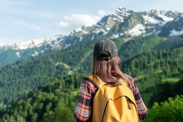 Naklejka premium Young woman with a yellow backpack stands in front of majestic mountains, surrounded by lush greenery, enjoying the breathtaking view of nature's beauty and tranquility