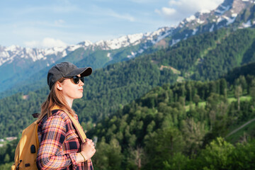 Young woman with backpack stands on mountain overlook, gazing at scenic landscape of lush green trees and snow-capped peaks under a bright blue sky, embodying adventure and exploration, copy space