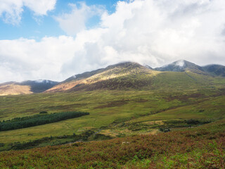 Green hills and mountains rise in the distance, partly covered by low-hanging clouds on Carrauntoohil walkway. Sunlight shines on the slopes in County Kerry, Ireland.