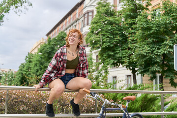 Young red haired woman sitting on a railing, smiling and looking away, with her bicycle next to her