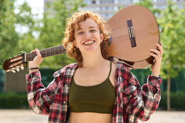 Portrait of cheerful redhead young woman musician holding acoustic guitar on shoulder smiling in...