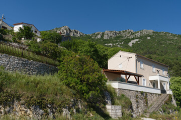 Houses cling to a lush green hillside, overlooking a stunning mountain range under a clear blue sky, evoking a sense of tranquility.