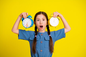 Surprised young girl holding two alarm clocks against a yellow background, expressing astonishment...