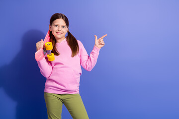 Smiling young girl in a pink hoodie holding a skateboard and gesturing playfully against a purple...