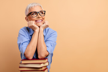 Delighted senior woman in glasses resting hands on a book stack against a beige backdrop,...