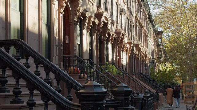 Two women walking on a sidewalk in front of an iconic row of brownstones in a residential district in Manhattan, New York City during autumn
