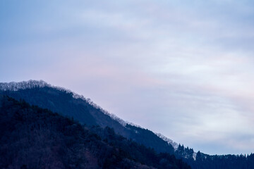 Fototapeta premium 埼玉県秩父の、冬の山の稜線と夕暮れの空。日没後の静寂の中、稜線に並ぶ木々が白く輝く、水墨画のように美しい風景。日本の自然が持つ、静かでミニマルな美しさを表現した一枚。