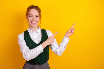 Happy student pointing with both hands in front of bright yellow background, wearing academic...