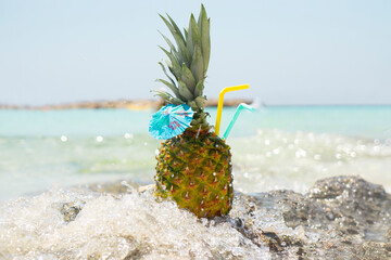 Fresh pineapple fruit cocktail with a straw and a blue umbrella stands on a rock in a spray of sea water on a tropical background. Summer vacation and summer vacation concept.