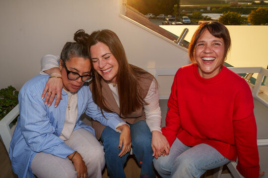 Three female friends are enjoying time together, laughing and embracing on a terrace at sunset
