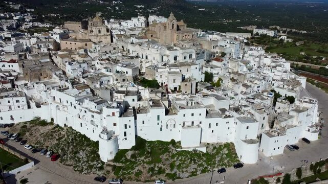 Aerial drone footage of Ostuni, a whitewashed hilltop town in Apulia, southern Italy, showing the historic old town, narrow streets, cathedral, and surrounding olive groves under clear skies.