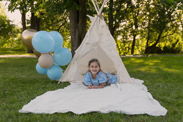 a girl is playing in her teepee in the garden, and blue and golden balloons are blowing in the wind