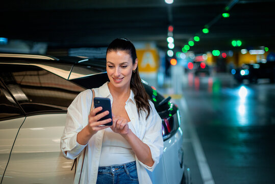 Brunette haired woman standing next to her car in car park and using phone