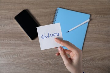 Woman holding paper note with word Welcome at wooden table, top view