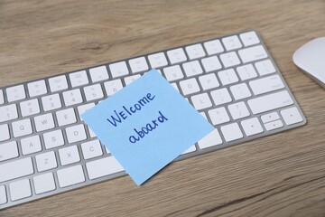 Sticky note with phrase Welcome Aboard, computer keyboard and mouse on wooden table, closeup