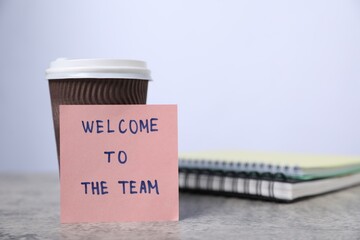 Sticky note with phrase Welcome To The Team, coffee and notebooks on grey table, closeup