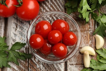Tasty pickled tomatoes in glass bowl, parsley and garlic cloves on wooden table, flat lay