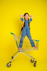 Cute young girl standing confidently in shopping cart against vibrant yellow background