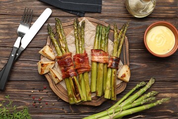 Tasty cooked asparagus served with bacon and sauce on wooden table, flat lay